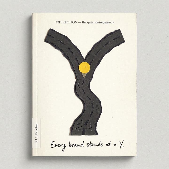 Manifesto Vol. 01 — a hand-cut paper road forking into a Y, with a single yellow dot at the fork, captioned “Every brand stands at a Y.”